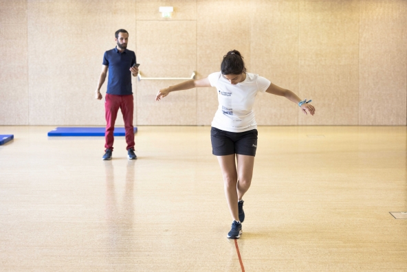 Eine Frau geht eine Linie entlang in einer Turnhalle, dahinter steht ein Mann, der auf sein Smartphone blickt
